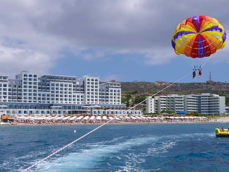 Resort sulla spiaggia con più edifici, parasailing sull'acqua e barca in primo piano.