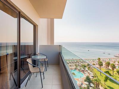 Balcone con tavolo, due sedie, ringhiera in vetro, vista sul mare e sul resort.