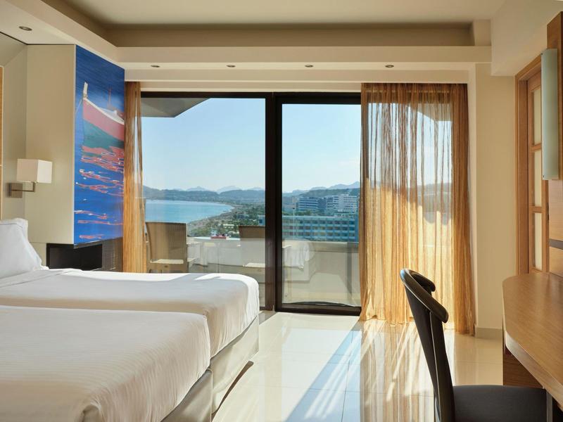 Camera d'hotel luminosa con due letti e balcone con vista mare e montagne.