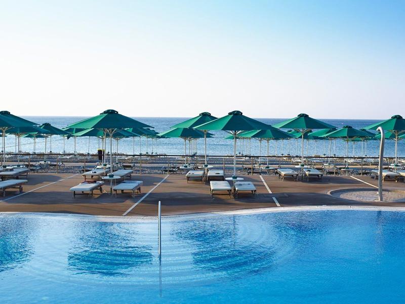 Vista di una piscina e lettini con ombrelloni verdi sulla spiaggia sotto un cielo limpido.