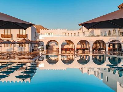 Großer Pool mit Wasserspiegelung vor weißem Gebäude mit Bögen, blauer Himmel, Sonnenschirme am Rand.