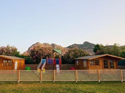 Zwei Holzhütten mit Spielplatz vor grüner Wiese und Bergen im Hintergrund.