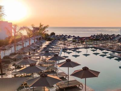 Großer Hotelpool mit Sonnenschirmen und Palmen bei Sonnenuntergang am Meer.