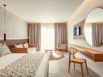 Bright hotel room with double bed, wooden chair, round mirror, and soft curtains over large window.