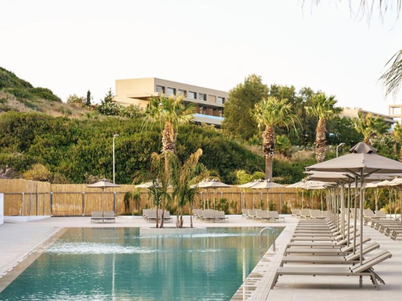 Modern pool area with sun loungers and palm trees in front of a hotel building on a hill.