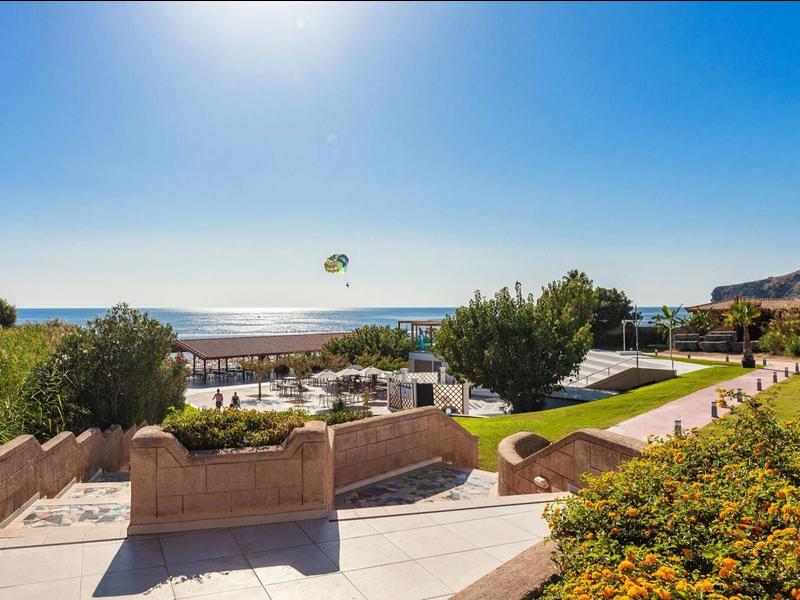 Seaside resort with parasailing, beach, pool, greenery and mountains under a sunny sky.