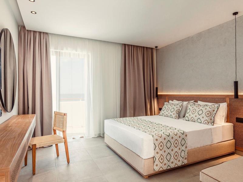 Bright hotel room with double bed, desk, chair, and large window with curtains.