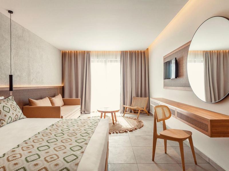 Bright hotel room with double bed, wooden chair, round mirror, and soft curtains over large window.