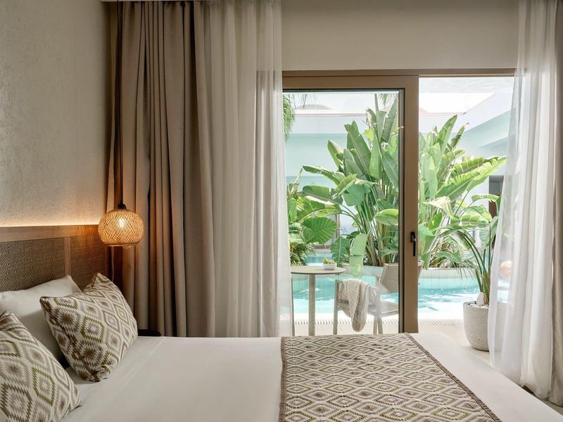 Modern hotel bedroom with double bed and sliding door overlooking pool and greenery.
