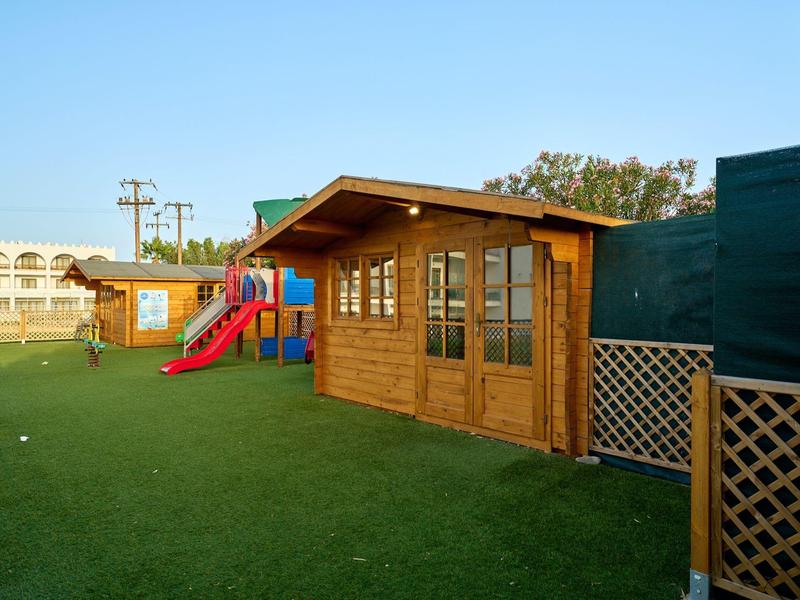Wooden cabins and a slide on a green lawn under clear sky at a hotel playground