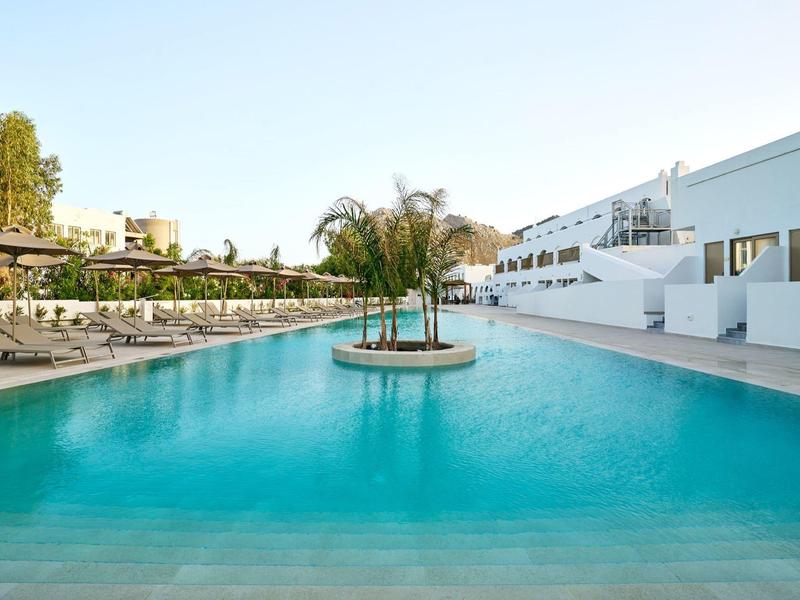 A large outdoor pool with an island of palm trees in the center, surrounded by sun loungers and white buildings.