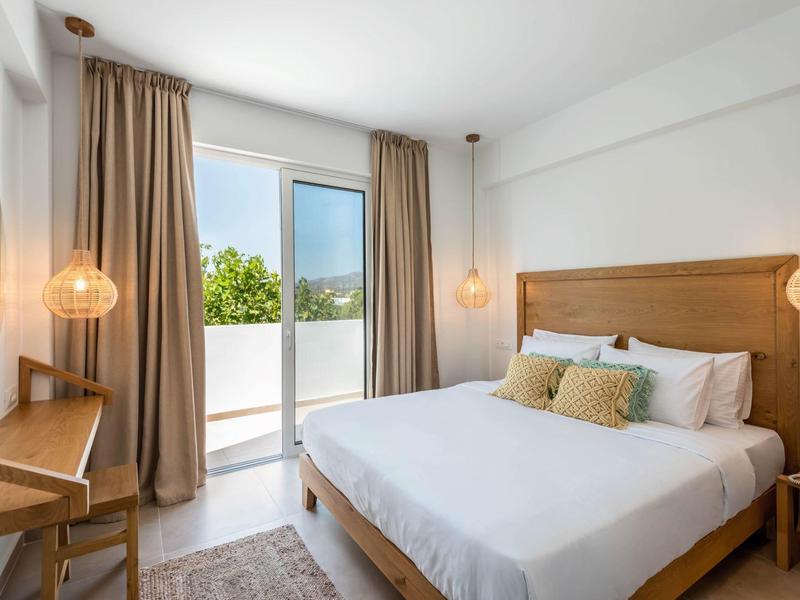 Bright hotel room with double bed, wooden furniture, and terrace overlooking green trees.