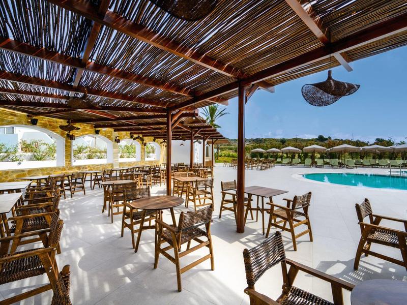 Covered outdoor seating area with chairs and tables beside a pool and resort buildings.