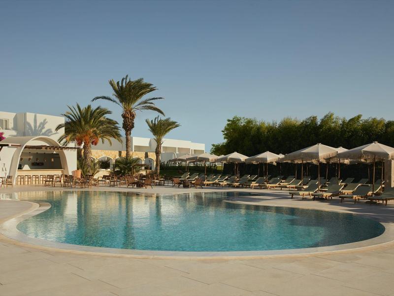 Round hotel pool with lounge chairs and umbrellas, surrounded by palm trees and clear blue sky.