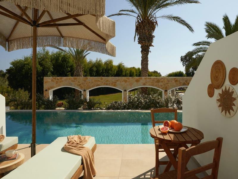 Hotel pool area with sun umbrella, lounge chair, and table with fruit, surrounded by palm trees.