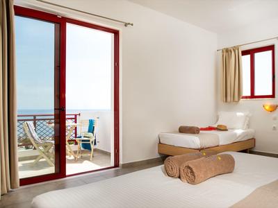 Světý pokoj v hotelu s balkonem a výhledem na moře, dvě postele a béžové stěny.