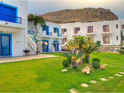 Jardín verde con palmeras frente a edificios blancos y azules y colinas al fondo.