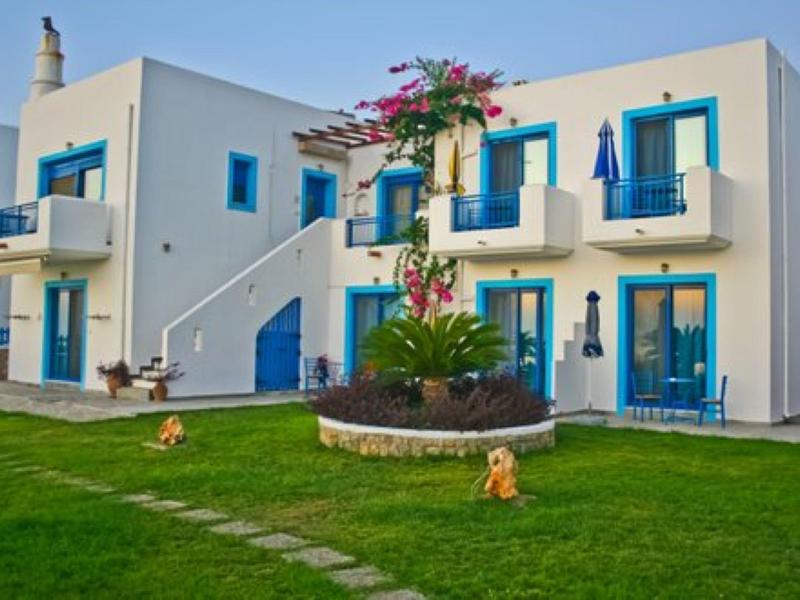 Bílý hotel s modrými dveřmi a okny, obklopený zeleným trávníkem a květinami.