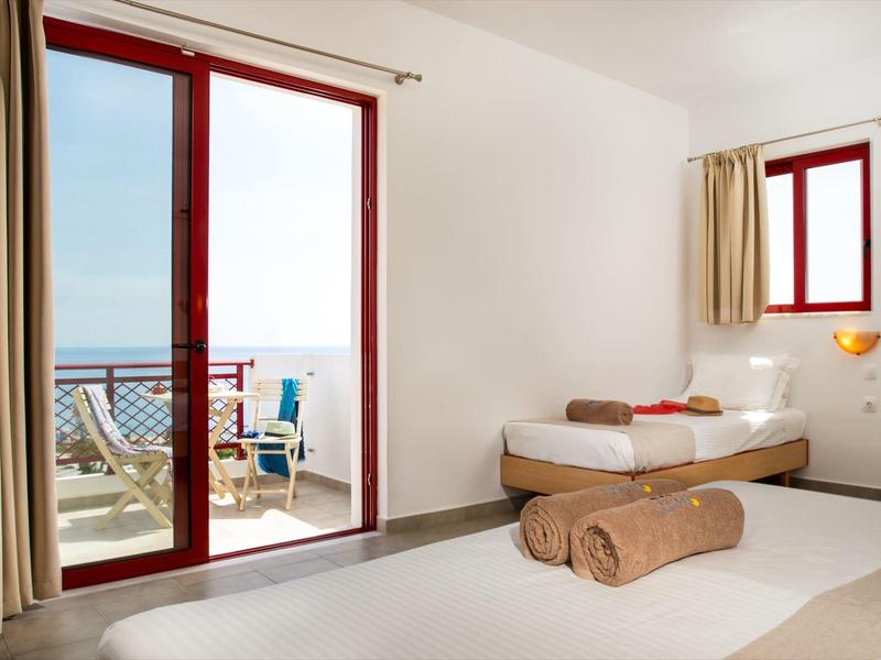Světý pokoj v hotelu s balkonem a výhledem na moře, dvě postele a béžové stěny.