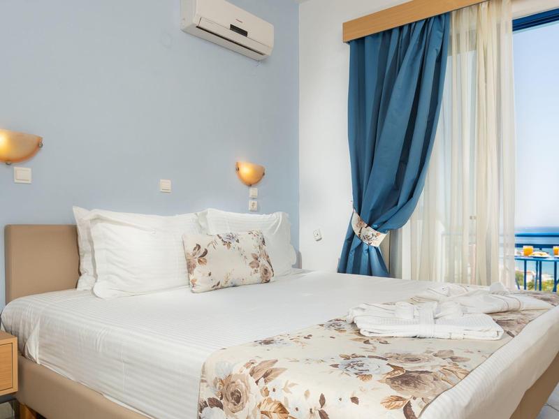 Světly hotelový pokoj s manželskou postelí, klimatizací a výhledem na moře oknem.