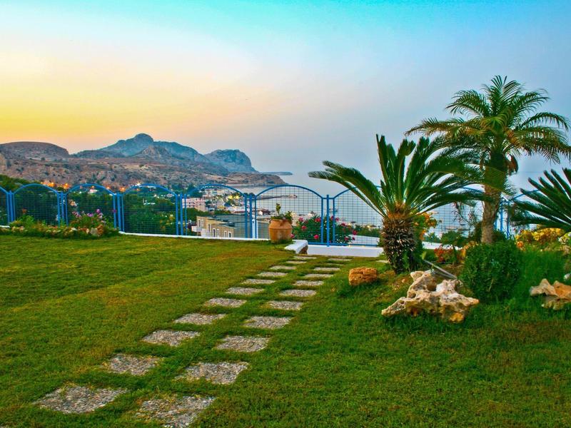 Zelená zahrada s kamennou cestou, palmami a výhledem na moře při západu slunce