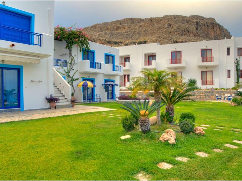 Hotelová budova s bílou fasádou a modrými akcenty, obklopená udržovanou zahradou s palmami.