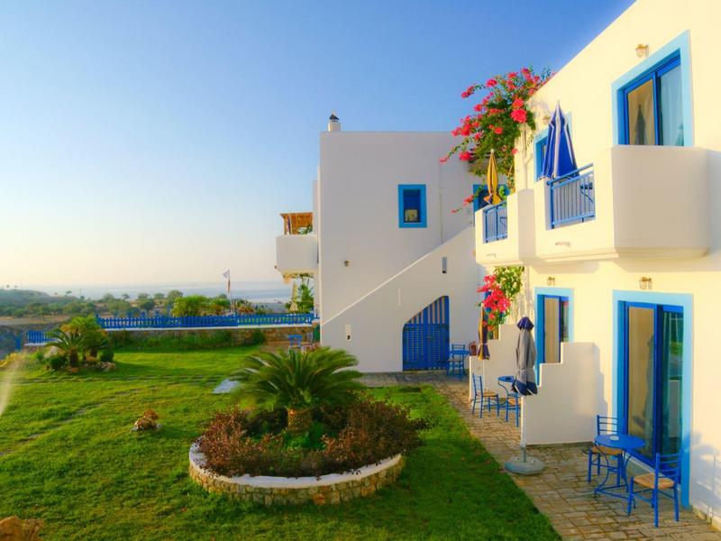 Bílá hotelová budova s modrými okenicemi, balkony a zelenou zahradou pod jasnou oblohou.