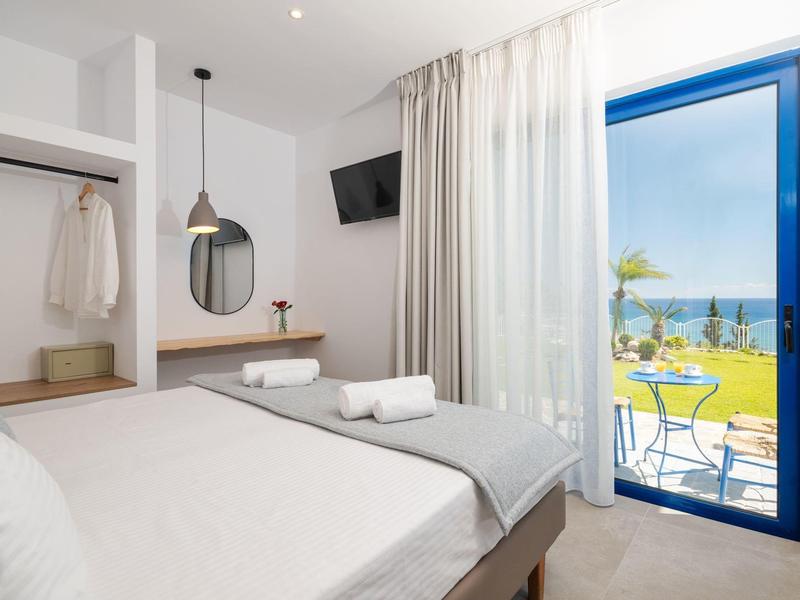 Světle hotelový pokoj s manželskou postelí, ručníky, balkonem a výhledem na moře.