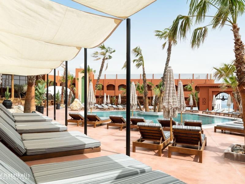 Area piscina dell'hotel con lettini, ombrelloni e palme alla luce del giorno.