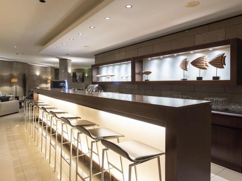 Nowoczesny bar hotelowy z długim białym blatem i wieloma stołkami barowymi pod ciepłym oświetleniem.