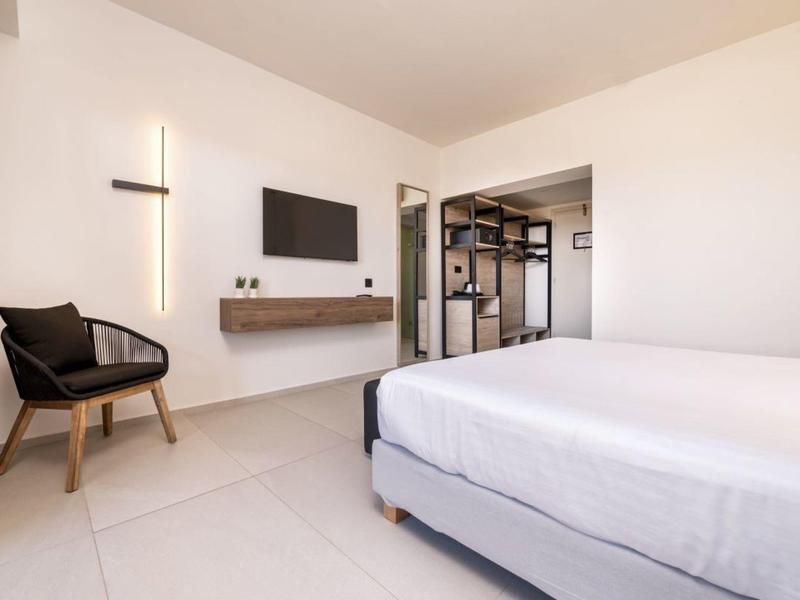 Nowoczesna sypialnia z podwójnym łóżkiem, telewizorem na ścianie, czarnym fotelem i otwartą szafą.