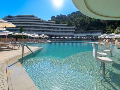 Großer blauer Pool mit weißen Barhockern und Sonnenschirmen vor modernem Hotelgebäude und Hügeln.