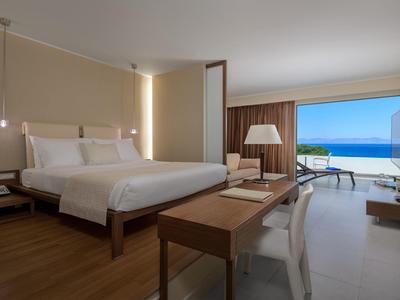Modernes Hotelzimmer mit großem Bett, Schreib­tisch, Sitz­ecke und Blick auf Meer und Terrasse.