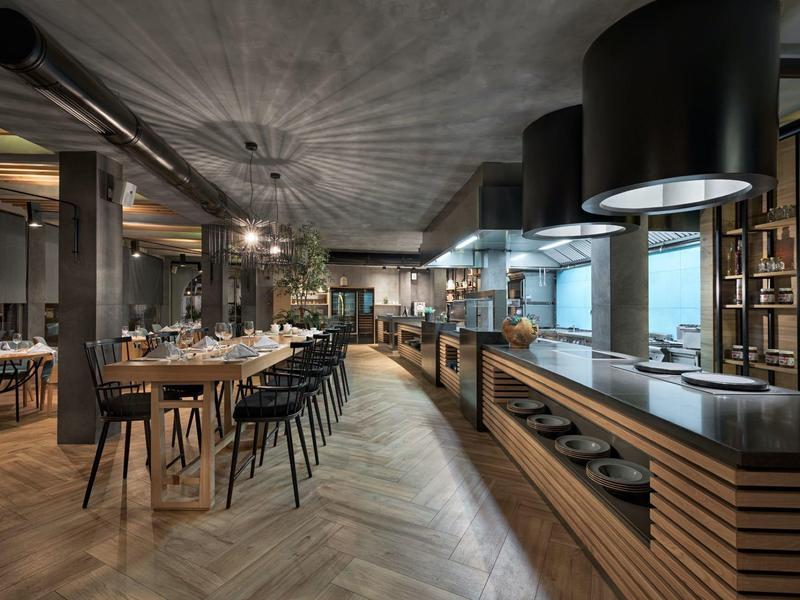 Modernes Restaurant mit Holzfußboden, langen Holztischen, schwarzen Stühlen und offener Küche.