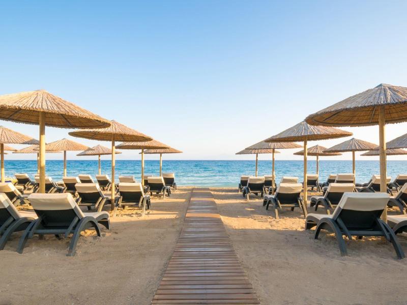 Strand mit Sand, Holzweg, Sonnenliegen und Strohhut-Sonnenschirmen unter klarem blauen Himmel.