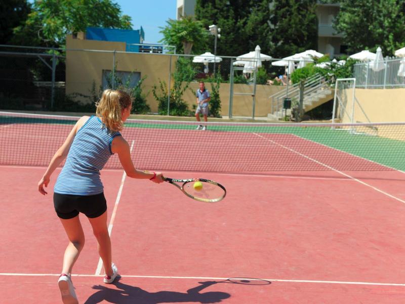 Frau spielt Tennis auf rotem Sandplatz bei sonnigem Wetter, umgeben von Bäumen und Gebäuden.