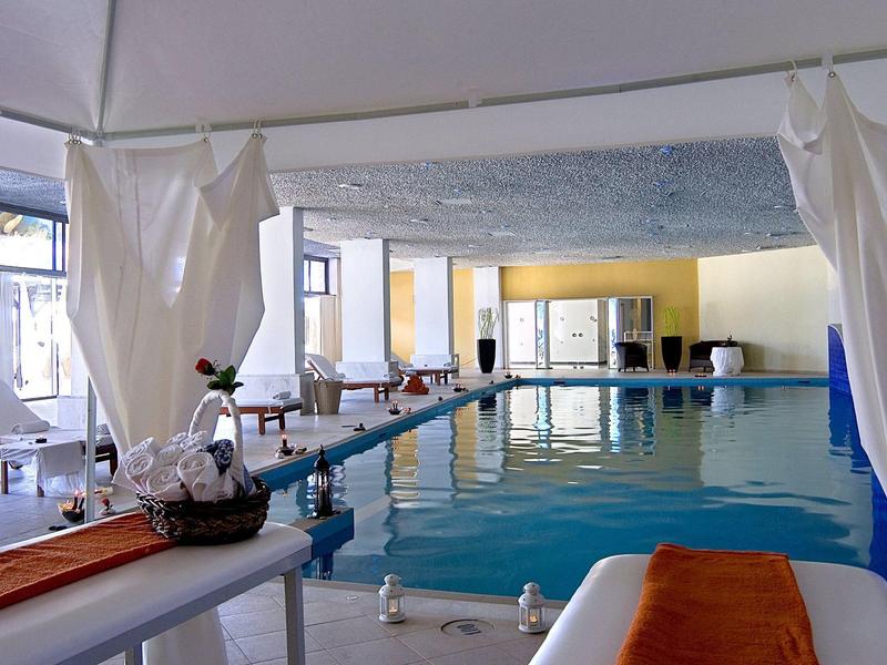 Innenraum eines Schwimmbads mit großem Becken, Sonnenschirmen und Liegen mit weißen und orangen Tüchern.