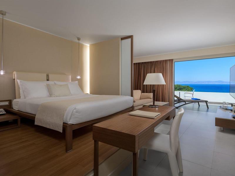 Modernes Hotelzimmer mit großem Bett, Schreib­tisch, Sitz­ecke und Blick auf Meer und Terrasse.