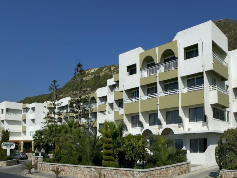 Immeuble d'hôtel blanc à plusieurs étages avec balcons devant une colline boisée et ciel bleu.