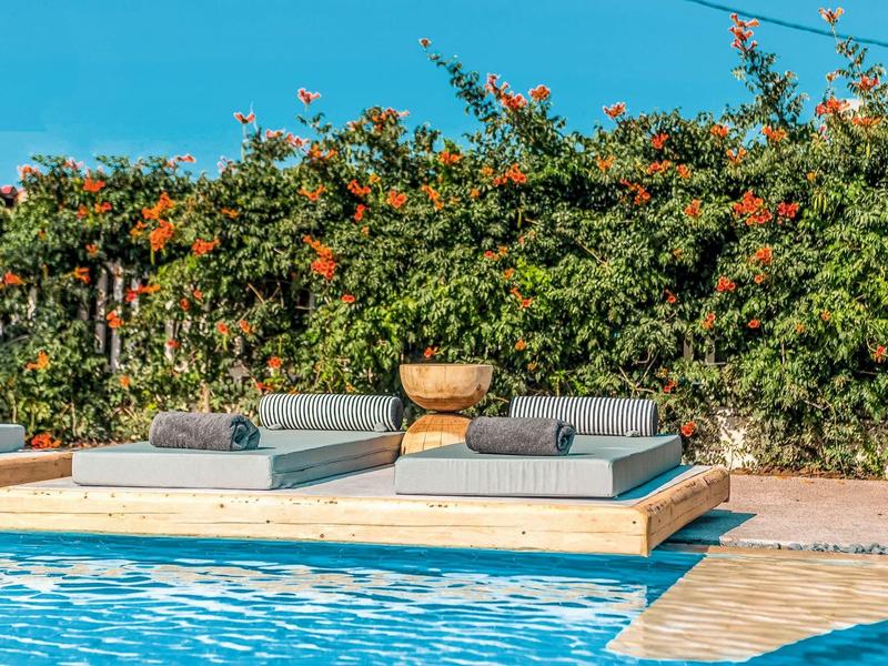 Piscina con terrazza in legno e lettini davanti a una siepe fiorita sotto un cielo azzurro.