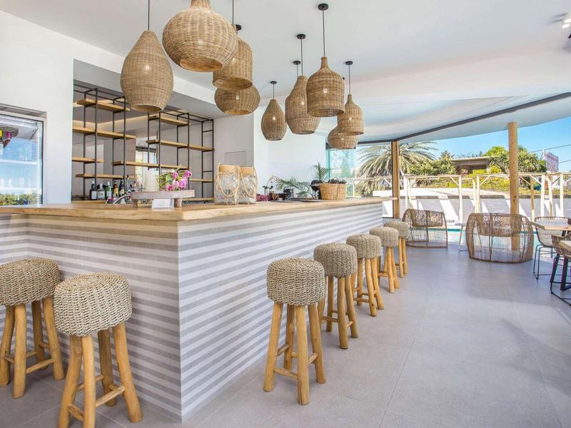 Bar luminosa con sgabelli in legno, lampade intrecciate e vista aperta su terrazza e piscina.