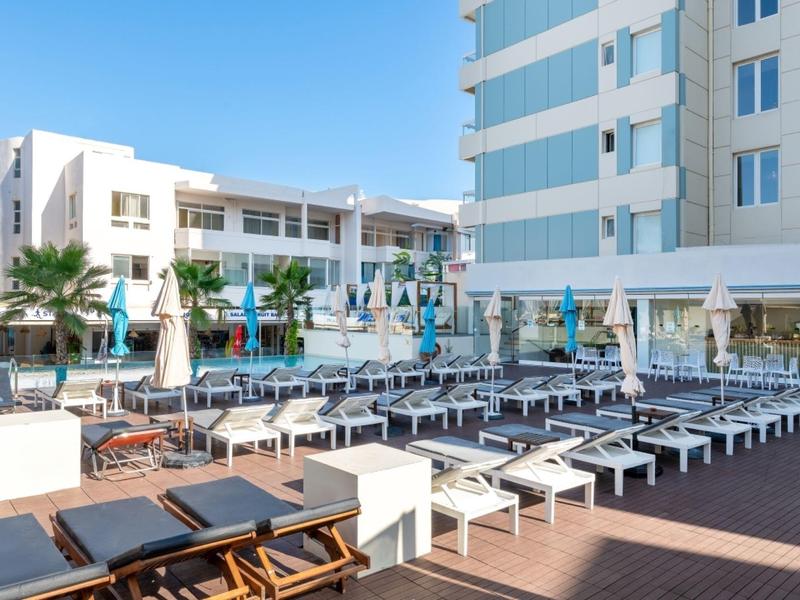 Veel ligstoelen met blauwe parasols bij een hotelzwembad onder een heldere hemel.