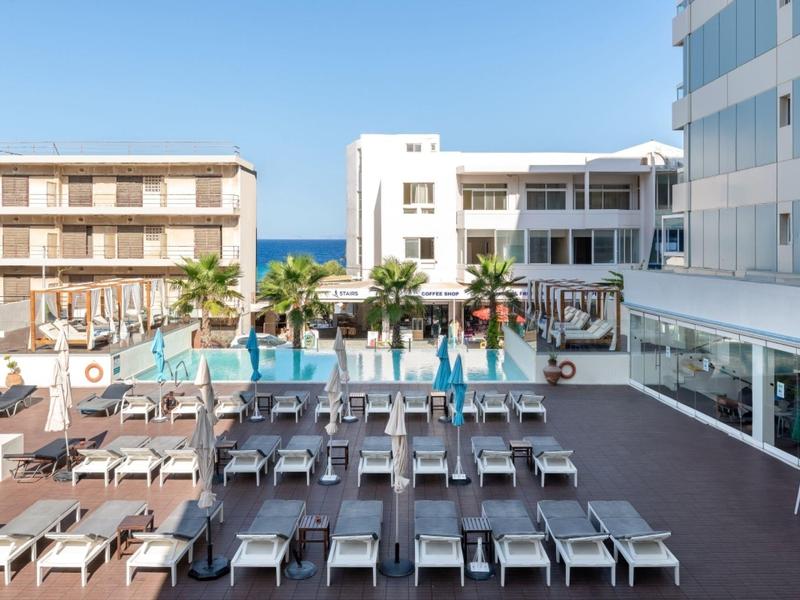 Uitzicht op een hotelzwembad met ligstoelen en moderne gebouwen op de achtergrond bij de zee.