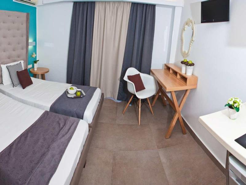 Modern hotel room with two single beds, desk, and kitchenette.