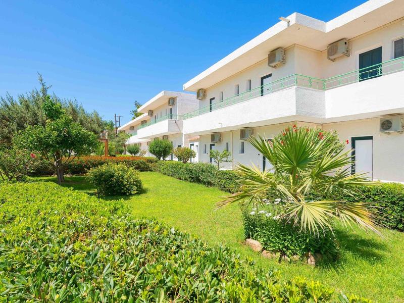 Modern white building complex with green gardens and palm trees under sunny weather.