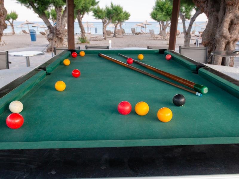 A green pool table outdoors with colorful balls under trees by the beach.