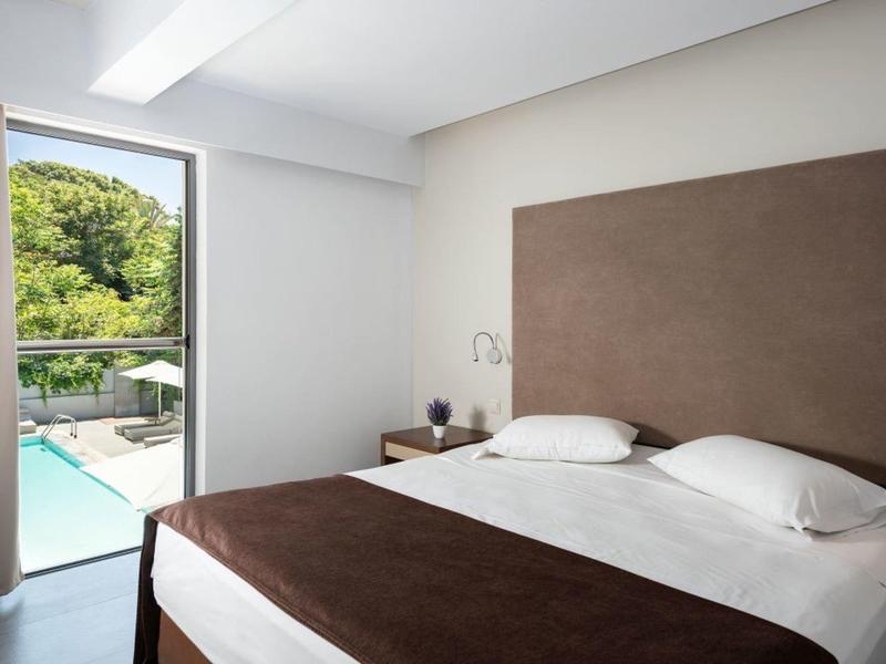 Bright hotel room with double bed, large headboard, and view of pool and green trees.