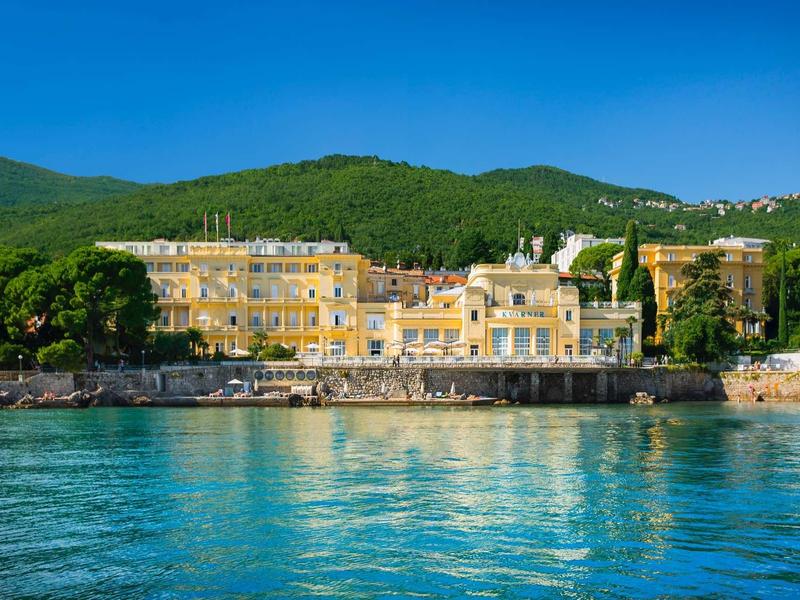 Duży żółty hotel nad brzegiem z zielonymi wzgórzami w tle i czystą, niebieską wodą.
