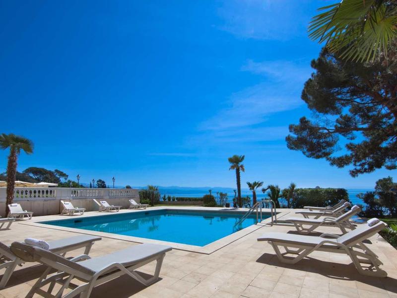 Strefa basenu z leżakami, niebieskie niebo, palmy i widok na morze w tle.