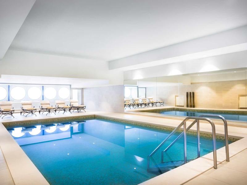 Jasny kryty basen z niebieską wodą i białymi leżakami na brzegach.